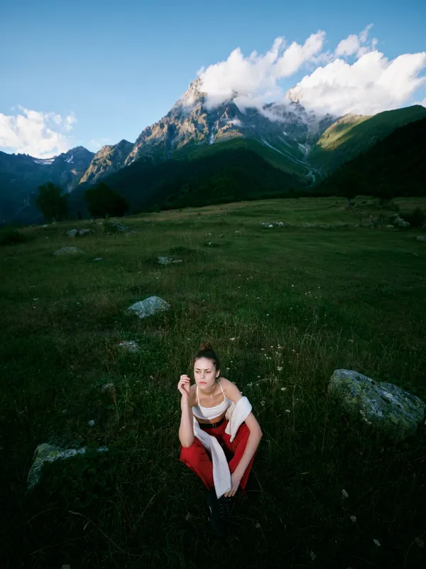 Woman kneels in a red dress on a meadow with rocks under a dramatic mountain range, capturing outdoor nature and a quiet landscape with soft clouds above