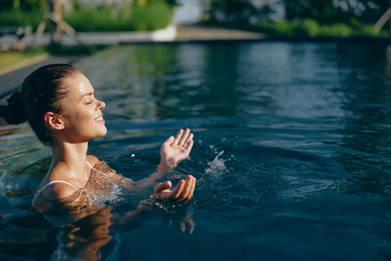 Young woman relaxing in outdoor swimming pool with eyes closed and hands raised. Concept of wellness, summer lifestyle, and peaceful moment in natural light.