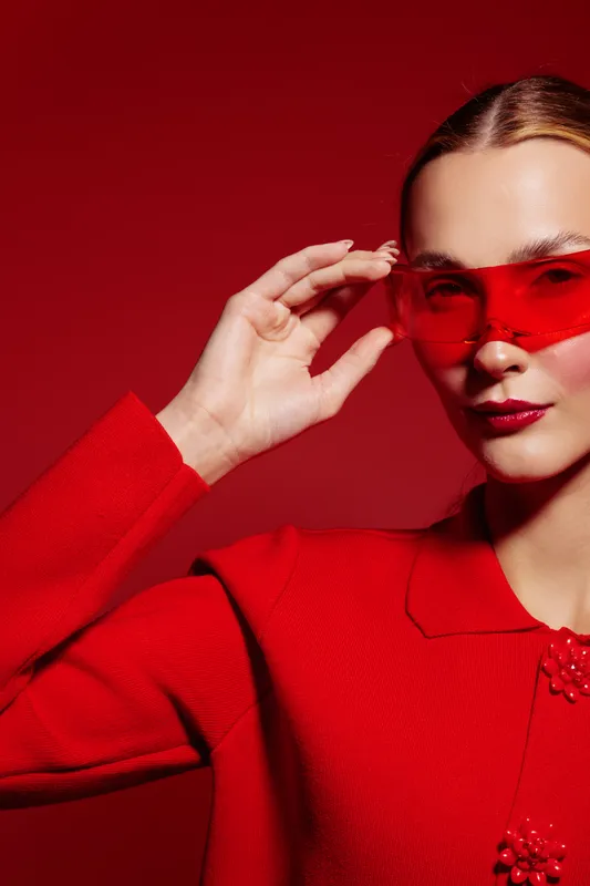 Stylish woman in red coat and red sunglasses poses against red background. Fashion portrait with glossy lips and sleek hair. Concept of modern style, elegance, and bold color contrast.