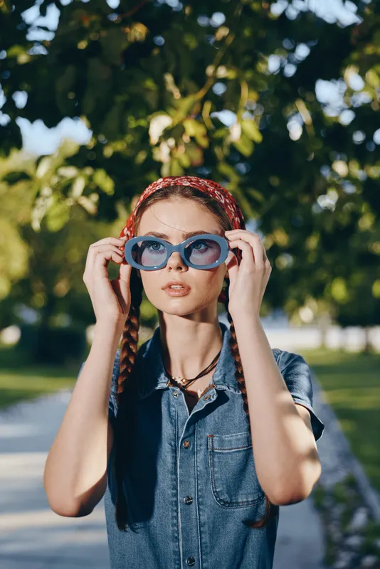 Lifestyle woman in modern boho-western denim street style adjusting blue sunglasses outdoors with natural light and film color effect, casual summer fashion vibe.