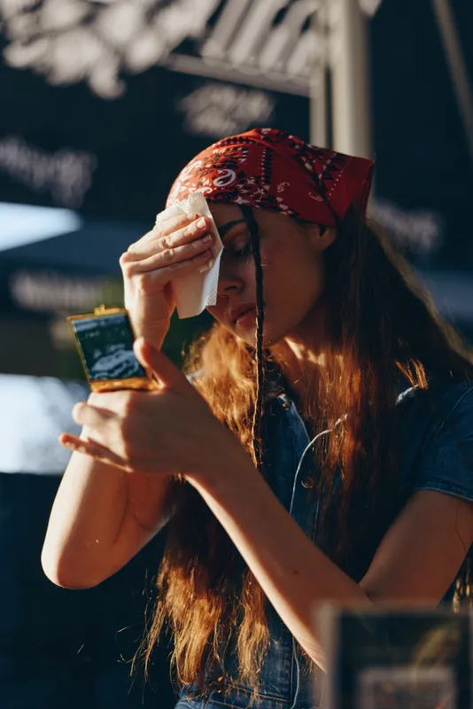 young woman wiping sweat with tissue outdoors wearing red bandana and casual denim top in sunny bright daylight