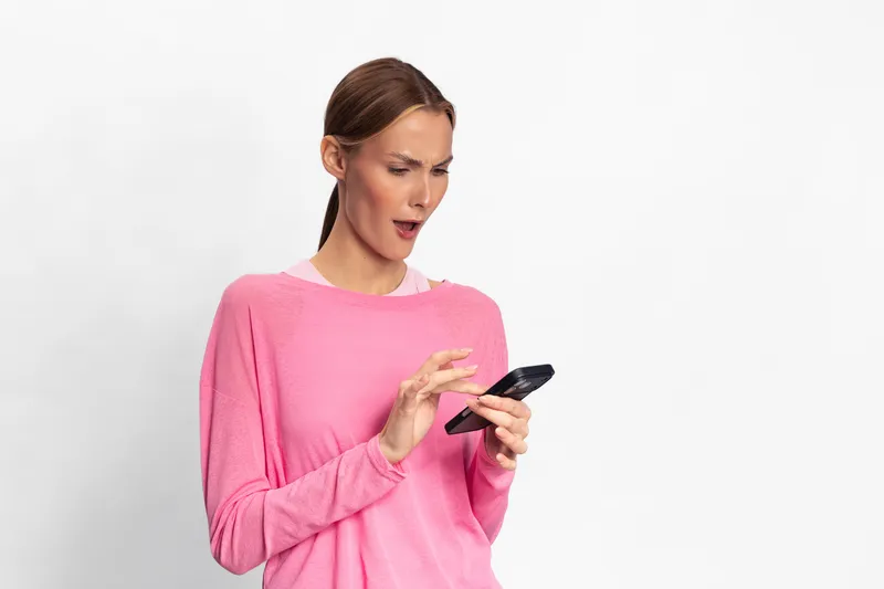 Surprised woman in pink sweater looks shocked at smartphone screen with raised eyebrows. Isolated on white background. Concept for unexpected news, message, mobile communication and emotion.