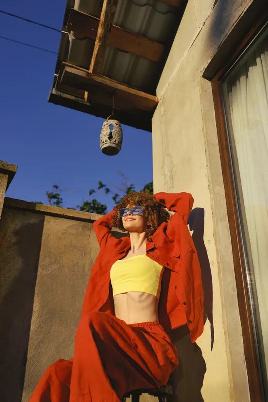 Woman lifestyle outdoor sunny clothing casual pose summer bright relaxed, a smiling woman wearing a yellow crop top and red jacket leans against a sunlit wall, conveying warmth and confident style