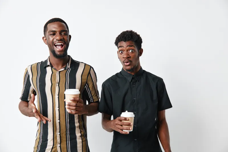 Two young black men with dark skin holding coffee cups and talking together in casual clothes on white background. Friends, communication and lifestyle with happy expressions.