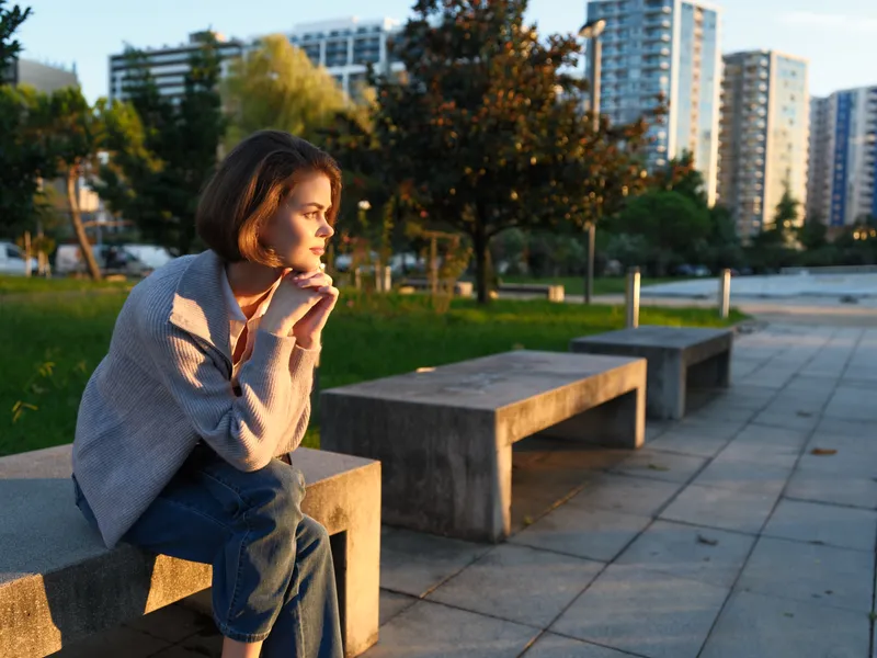 An attentive woman sits on a concrete bench in a sunlit urban park, her gaze distant and thoughtful, exuding calm concentration and a moment of personal reflection amid the city bustle.