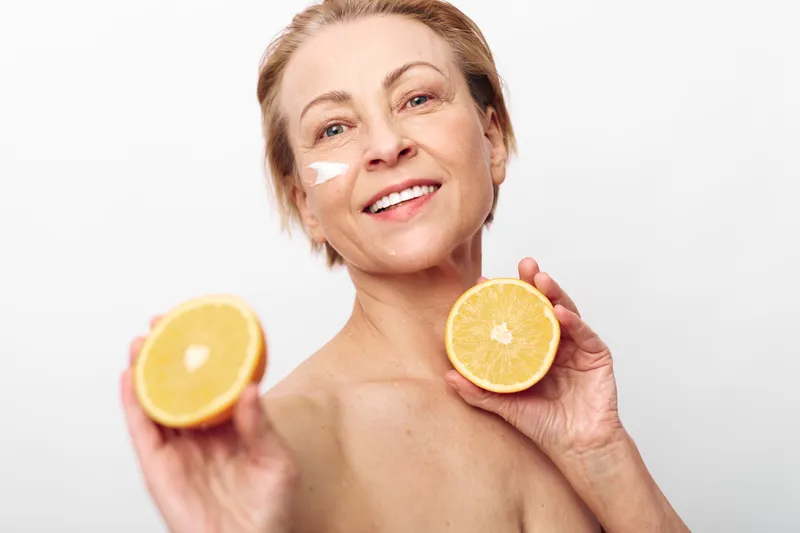 Smiling mature woman with natural skin holds fresh orange halves, applying white cream on cheek. Concept of healthy skincare, vitamin C, beauty, wellness, and natural cosmetic care.