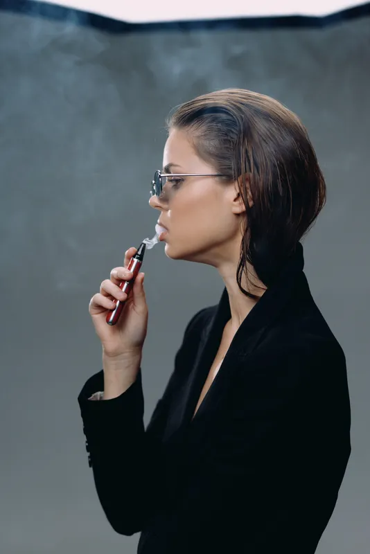 Stylish woman with glasses applying lipstick in a dark elegant outfit with modern hairstyle against gray background.