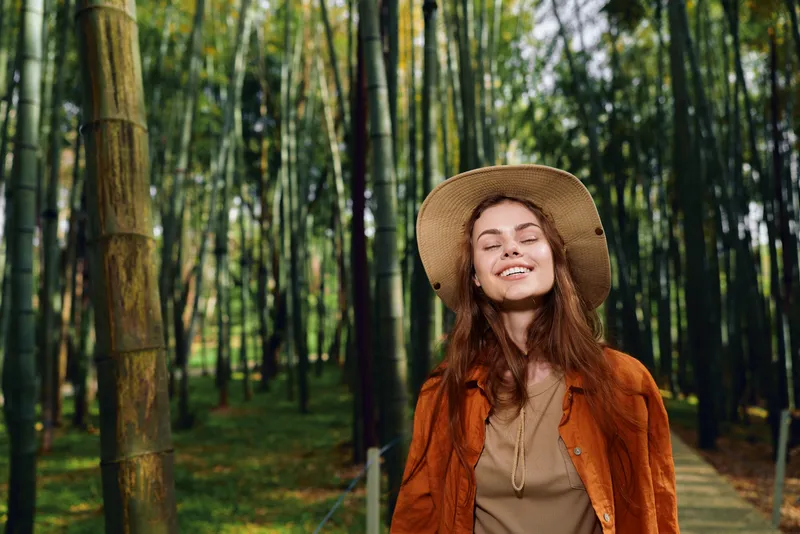 Woman in bamboo forest smiling with eyes closed wearing straw hat and brown jacket on a woodland path, peaceful portrait of nature walk and relaxed outdoor travel mood.