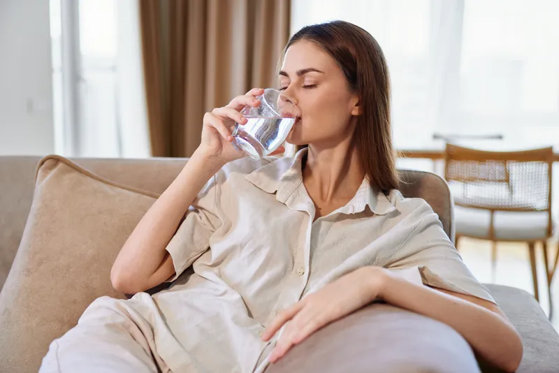 Woman drinking water from a glass on sofa at home, relaxed portrait of a young adult staying hydrated in living room, casual loungewear and natural daylight domestic scene