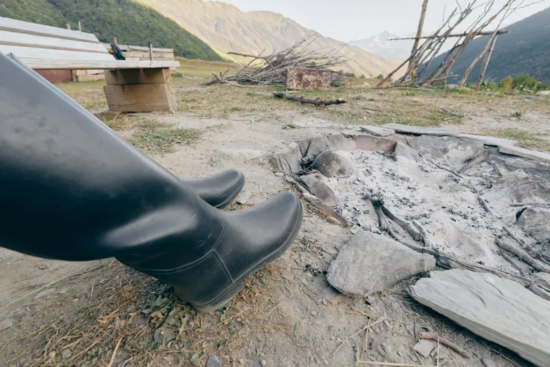 boots by a campfire ash at a cold firepit in mountains, outdoor resting scene with rubber footwear near burnt wood, rustic bench and valley view for hiking or camping break