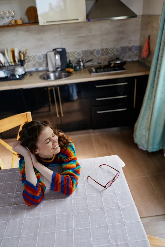 Cheerful woman indoors, leaning on a table in a bright kitchen, colorful sweater, warm light, candid moment, relaxed pose, cozy atmosphere, everyday living