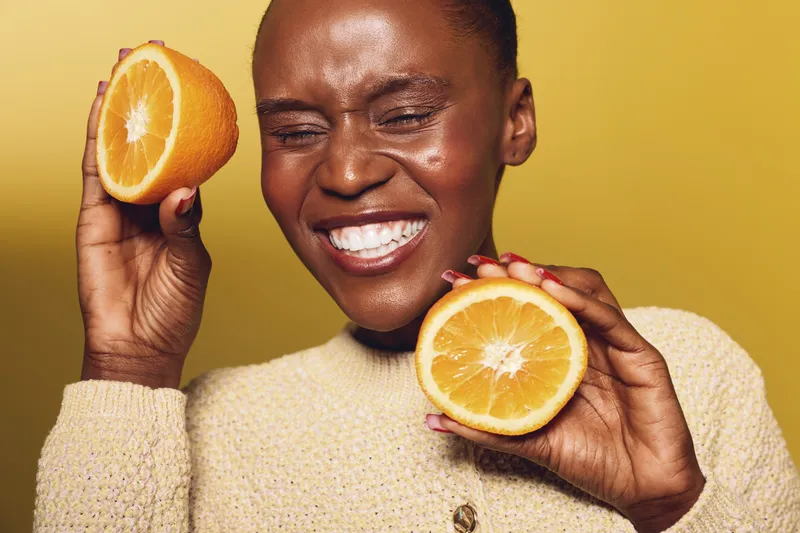 Smiling African woman with dark skin holds two orange halves near her face on yellow background. Concept of healthy lifestyle, vitamin C, fresh fruit, and natural beauty care.