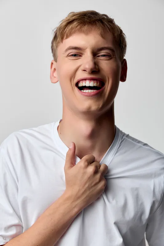 Smiling man laughing and tugging at a tshirt, conveying joy, energy and casual confidence for lifestyle, fashion and upbeat moments in a bright studio setting, ideal for advertising and social media