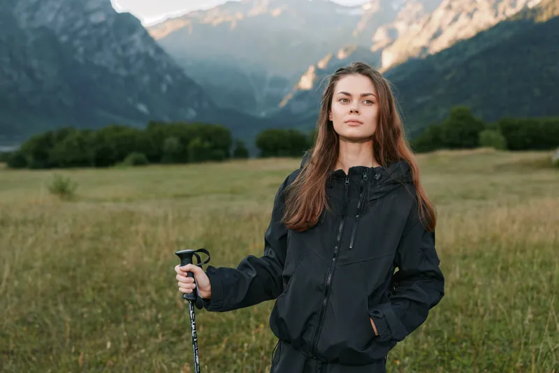 A poised woman stands outdoors in a wide valley with mountains in the distance. She wears a dark jacket and grips a trekking pole, ready for adventure in nature.