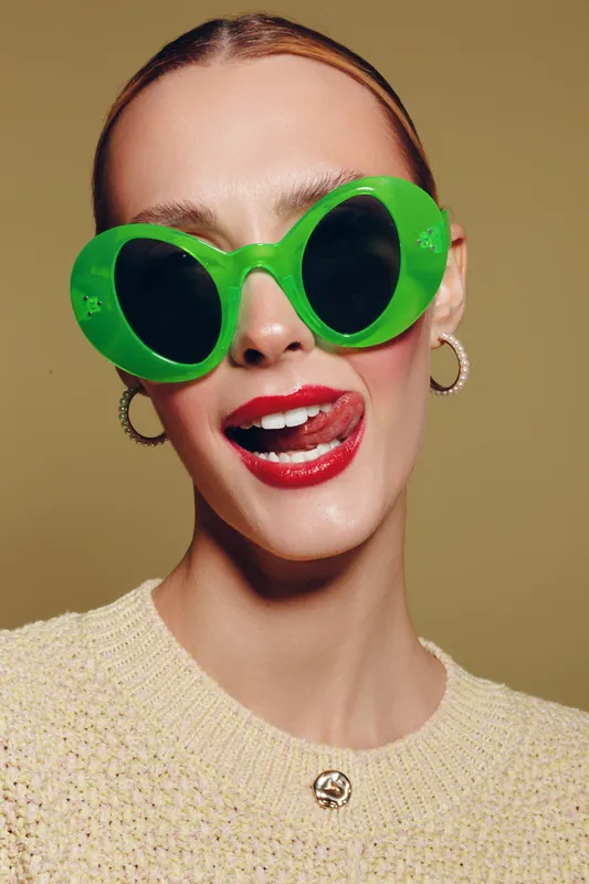 Young woman with slicked hair wears oversized green sunglasses and red lipstick, sticking out her tongue. Studio portrait on beige background with playful and bold fashion concept.