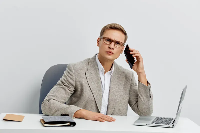Professional man in business suit wearing glasses sit at desk in modern office talking on mobile phone with serious expression. Workplace, corporate, communication, professional, workspace.