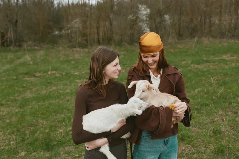 Two young women enjoy holding baby goats outdoors in a grassy field during spring, sharing a joyful moment of connection and relaxation in casual clothing.