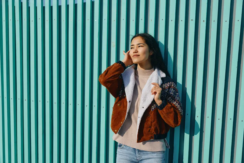 Stylish female model leaning against a turquoise striped wall in a casual outfit, shearling jacket over sweater, blue jeans, relaxed street style on a sunny day, backdrop of city.