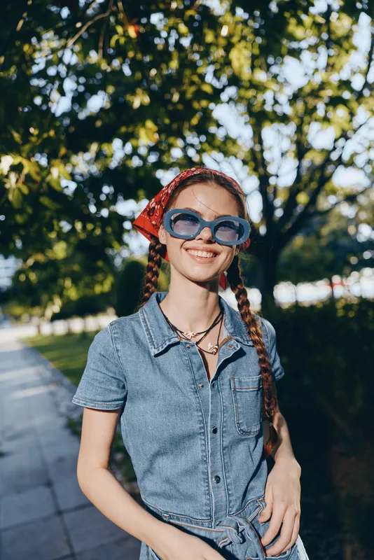 lifestyle woman wearing modern boho-western denim outfit and blue sunglasses smiles outdoors on sunny street, film color style with vibrant green background