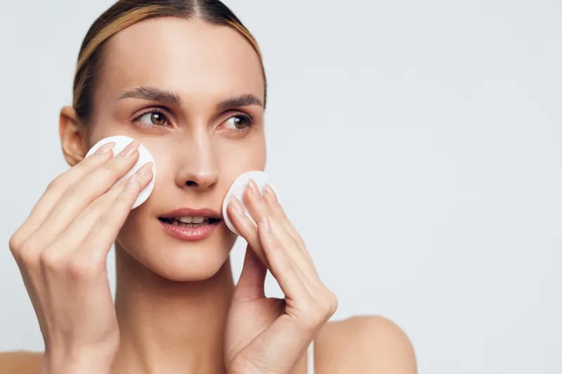 Smiling woman with clear skin uses cotton pads on her cheeks, close-up on a soft background. Concept of skincare, cleansing, and self-care. Gentle lighting creates a calm, fresh mood.
