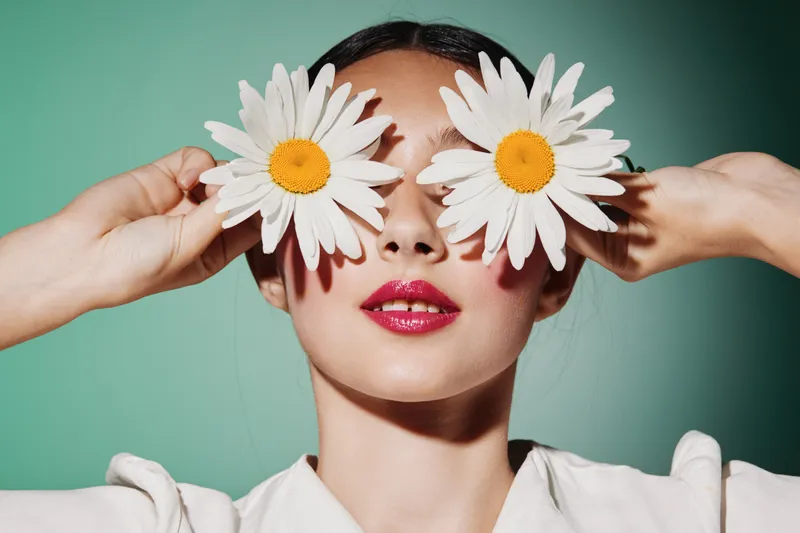 Young woman with fresh makeup holds two large white daisies over her eyes. Vibrant with glossy lips and smooth skin on a soft green background. Beauty creative and fashion concept.