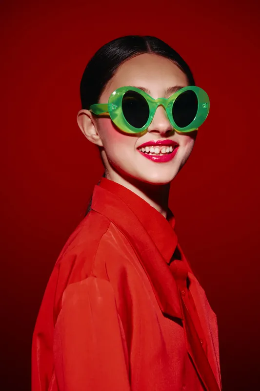 Portrait of a stylish young woman wearing striking green sunglasses and a vibrant red jacket against a red background in a beauty fashion creative concept.