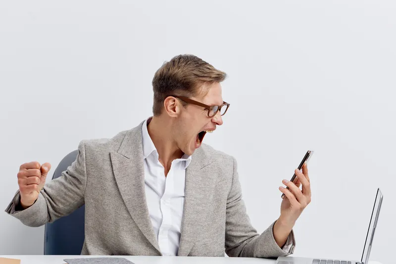 Confident young man wearing glasses and a gray blazer sitting at desk, looking at mobile phone with excitement and fist pump gesture, in bright modern office, emphasizing success and achievement conce