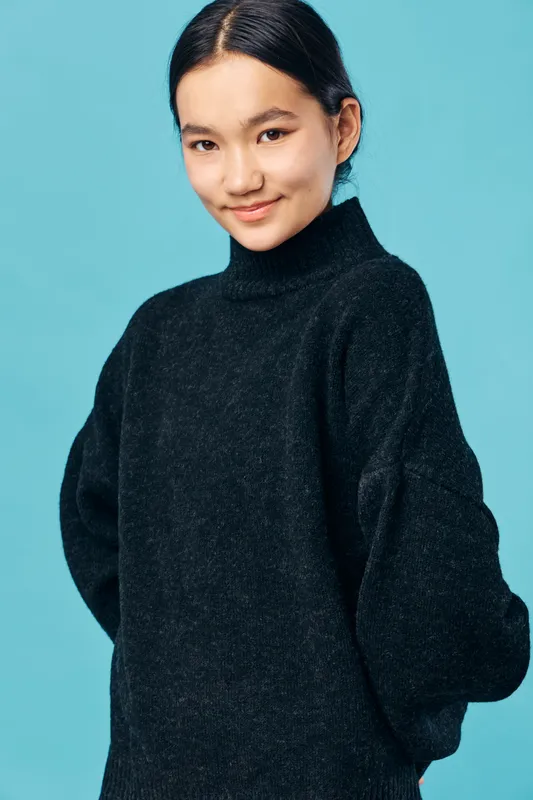 Young woman posing in black sweater with rolled sleeves against blue background. Casual style, confident smile, portrait with simple and clean composition in studio light.