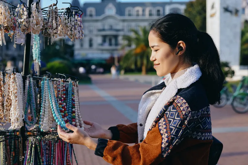 jewelry, necklaces, bracelets, accessories, shopping, street, market, vendor A shopper inspects beadwork at an outdoor stall, sunlight highlighting colorful beads and casual fashion in a vibrant plaza