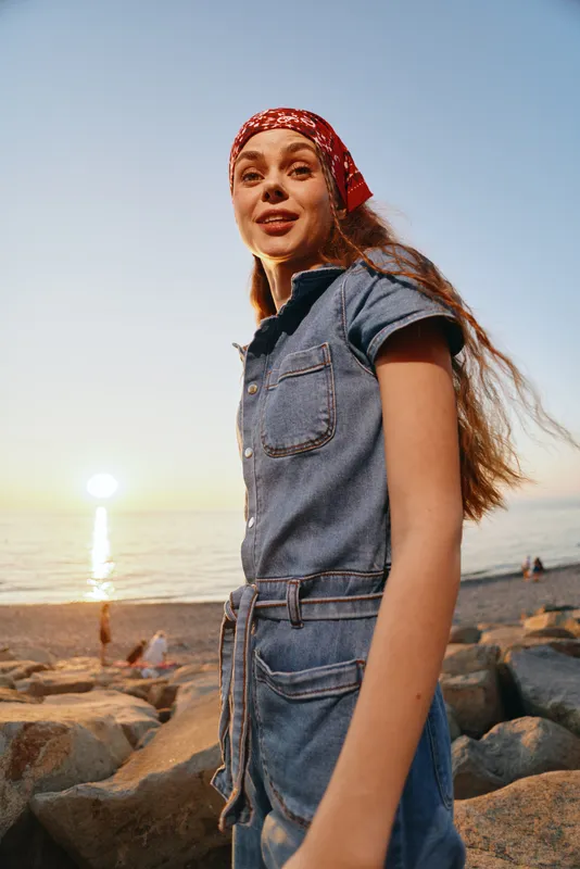 lifestyle woman in modern boho-western denim street style with red bandana standing on rocky beach at sunset, warm film color tones and relaxed vibe