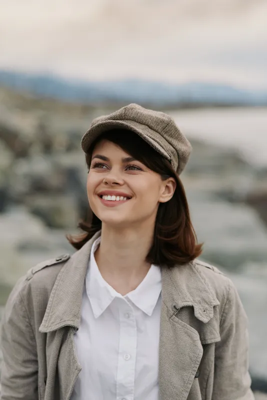 Young woman wearing a hat and coat smiles outdoors near water and rocks. Natural light highlights her joyful expression and casual autumn outfit. Concept of happiness and nature.