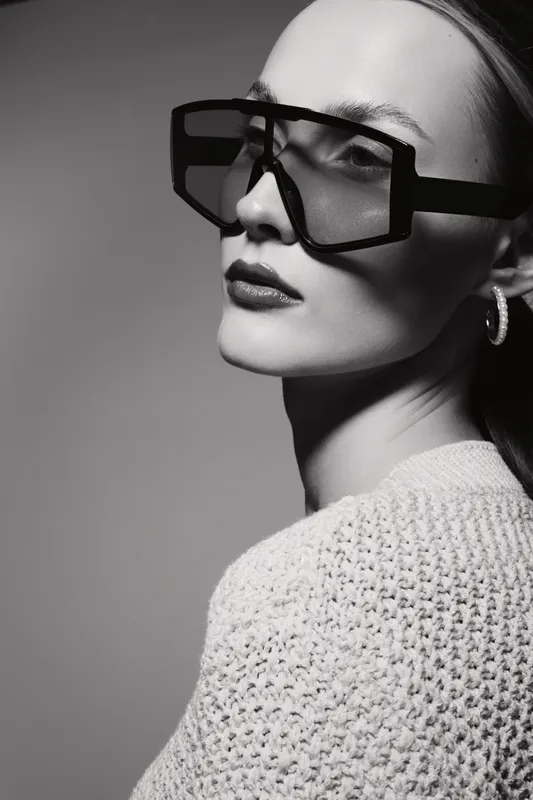 Stylish white woman with fair skin wearing oversized geometric sunglasses in black and textured sweater. Studio portrait in black and white.