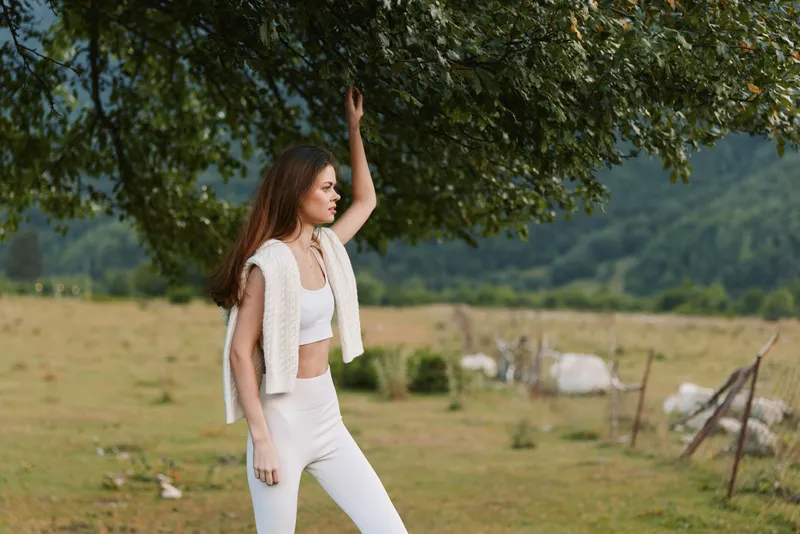 Young woman in white attire stands under a leafy tree in a rural field, creating a calm, relaxed lifestyle moment as distant cows graze nearby.