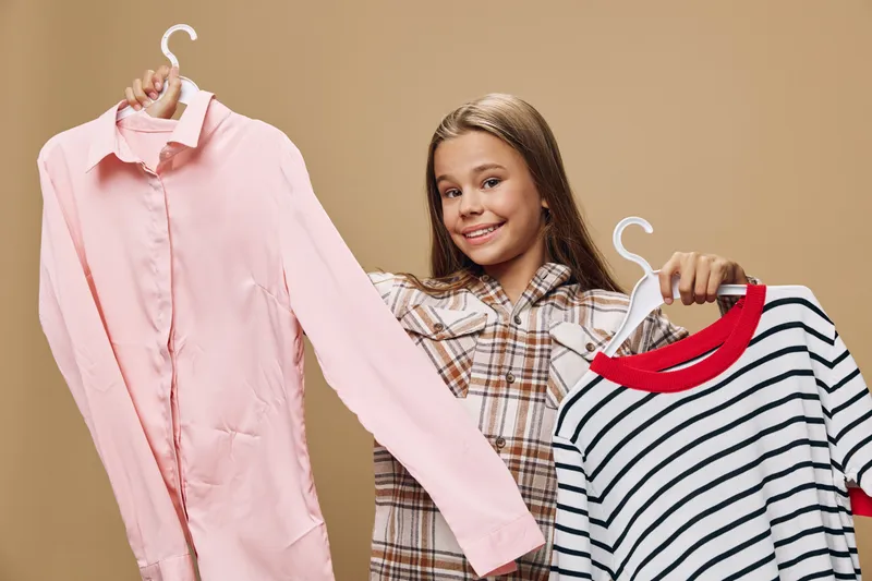 Girl holding two shirts on hangers smiling, comparing clothes with casual outfit against beige background in fashion shopping or wardrobe selection concept.