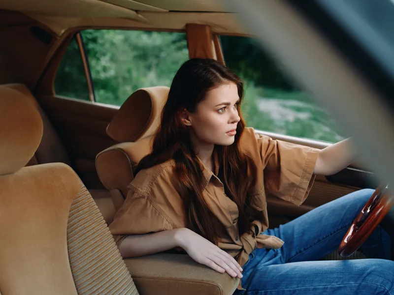 A thoughtful young woman wearing a casual brown shirt and blue jeans, gazing outside from the driver's seat of a vintage car amidst a lush green background.