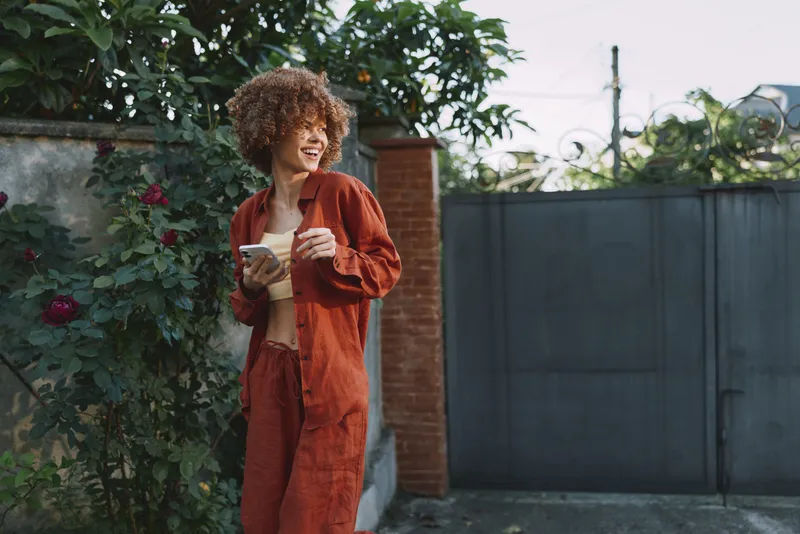 Young woman with curly hair in an orange jumpsuit stands in a garden, reading a notebook and smiling softly with a calm focused mood