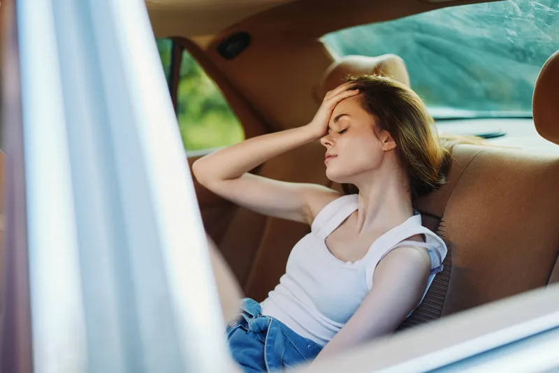 Young woman feeling stressed in the backseat of a car, holding her forehead, wearing casual clothes, with a blurred green background, reflecting emotions and modern lifestyle.