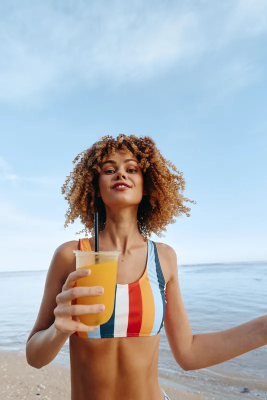 Young woman with curly hair wears a striped bikini and holds a glass of orange juice on a sunny beach, smiling softly as the sea and blue sky frame her carefree pose.
