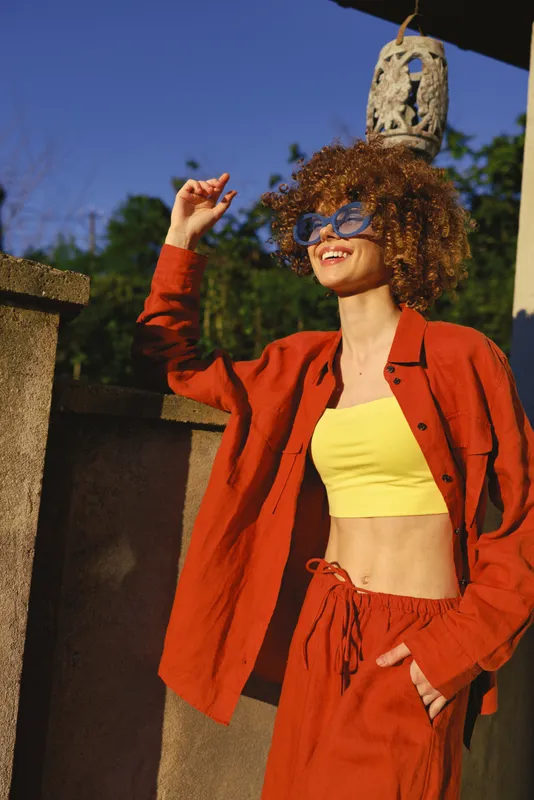 Cheerful woman outdoors in a vibrant orange outfit, smiling with sunny vibe, relaxed pose, bright daylight, natural colors, lush greenery background