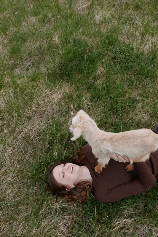 A woman lies on grass with a young goat perched on her chest, smiling in a sunlit field. An intimate lifestyle moment highlighting playful bond, calm happiness, and animal companionship in nature.