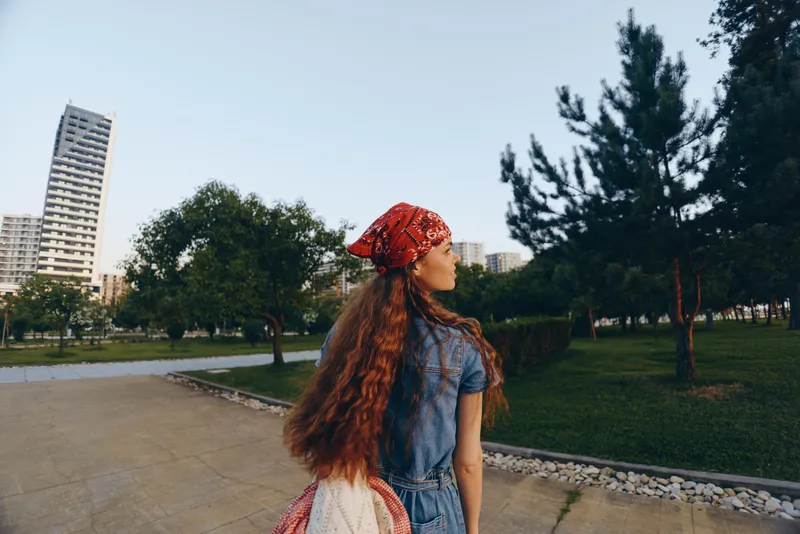 lifestyle woman wearing modern boho-western denim street style with long hair and red bandana walking outdoors in urban park under clear sky, film color aesthetic