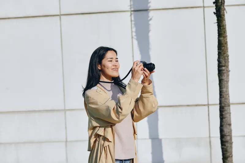 Woman in a beige coat holds a camera, capturing a sunny outdoor moment beside a pale wall and palm, emphasizing casual photography and everyday life