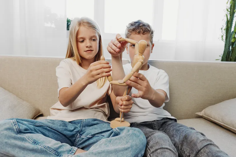 Two children sitting on a sofa playing with a wooden mannequin toy, focused on adjusting its limbs in a bright living room with natural light. Concept of creativity and childhood.
