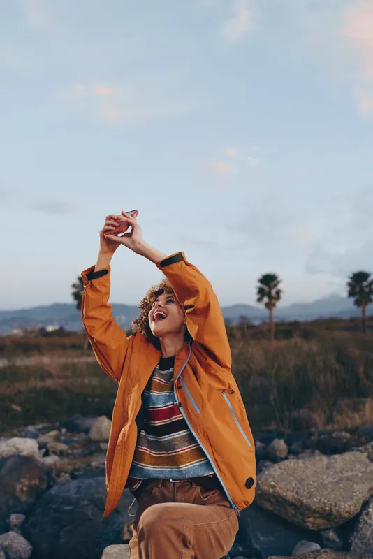 woman lifestyle portrait outdoors in orange jacket with arms raised toward sky over mountains, warm light, happiness, joyful relaxed expression, adventure moment, fresh air mood