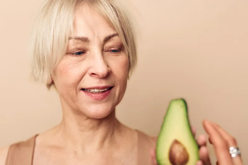 Smiling mature woman with short blond hair holds fresh halved avocado in hand, looking at fruit with interest. Healthy lifestyle concept, natural food, wellness, and nutrition focus in soft light.