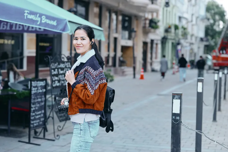 Young woman wearing a patterned fleece jacket and jeans models streetwear style in an urban shopping district, carrying a backpack, enjoying a casual city stroll