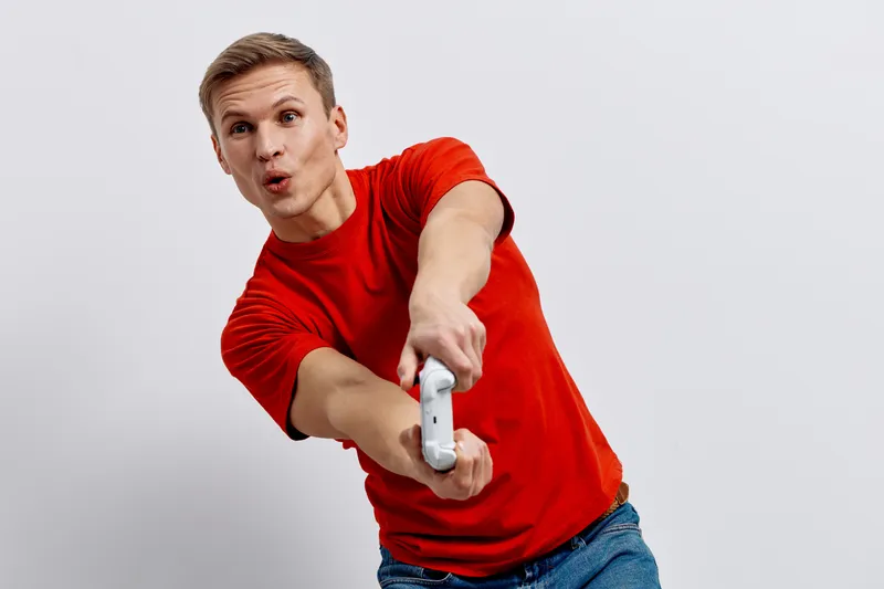 Excited young man in red t-shirt holding remote control, making funny face and pointing forward, on plain white background, studio portrait, lifestyle concept, expressive and energetic mood.