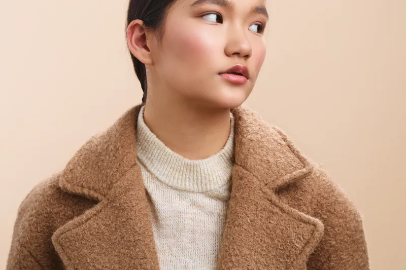 Asian woman in a cozy beige coat gazes off to the side, showcasing soft lighting, warm tones, and a gentle expression that conveys calm confidence and style.