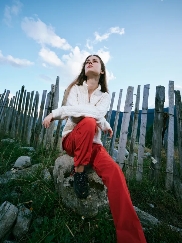Outdoor fashion scene with a stylish figure perched on a rock beside a weathered fence, wearing a white sweater and red pants, conveying casual confidence and natural charm.