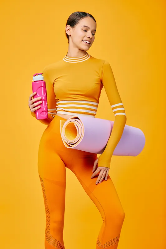 Vivid fitness scene with a confident woman in bright orange activewear, holding a water bottle and rolled yoga mat against a sunny yellow backdrop, ready for a strong workout and healthy lifestyle.